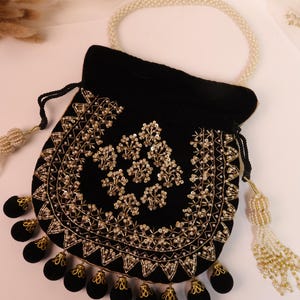 May include: A black velvet pouch with gold embroidery and bead detailing. The bag has a pearl handle and decorative tassels. The design features floral patterns and a scalloped edge, creating an elegant accessory.