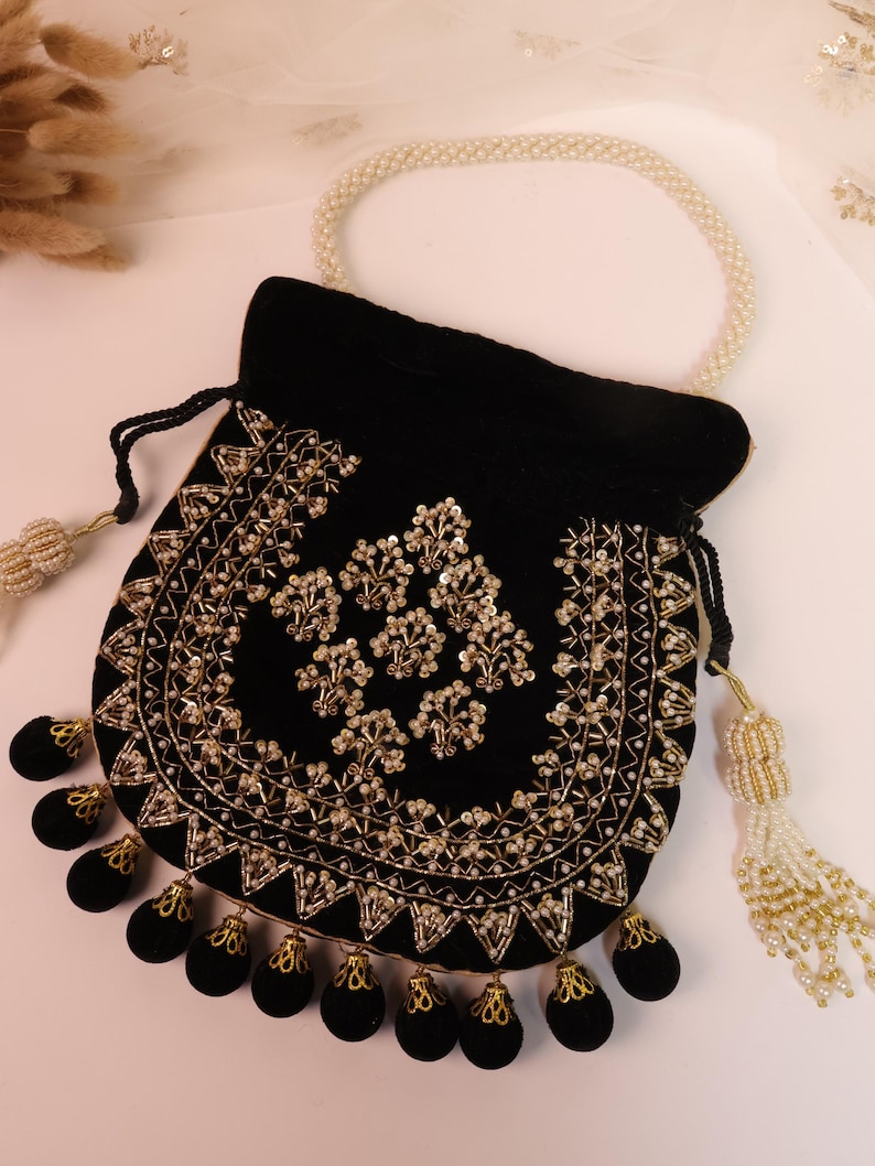May include: A black velvet pouch with gold embroidery and bead detailing. The bag has a pearl handle and decorative tassels. The design features floral patterns and a scalloped edge, creating an elegant accessory.