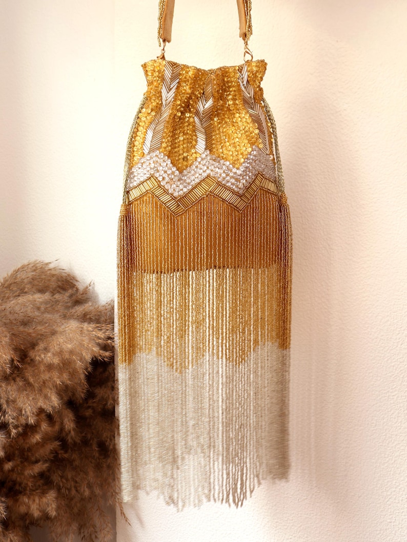 May include: A drawstring pouch bag featuring intricate gold and silver beadwork. The upper section displays a chevron pattern of gold and silver beads, while the lower portion consists of long, flowing gold and silver beaded fringe tassels.