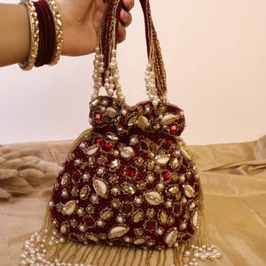 May include: A burgundy beaded handbag with a rounded shape, adorned with pearls, gold accents, and small seashells. The bag features a beaded handle and a fringe of pearls along the bottom. Gold and burgundy bracelets are visible on the wrist.