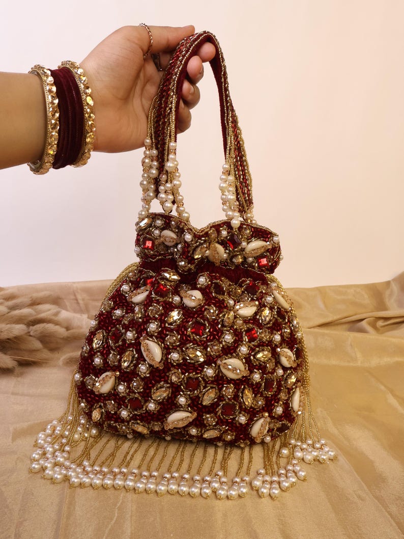 May include: A burgundy beaded handbag with a rounded shape, adorned with pearls, gold accents, and small seashells. The bag features a beaded handle and a fringe of pearls along the bottom. Gold and burgundy bracelets are visible on the wrist.