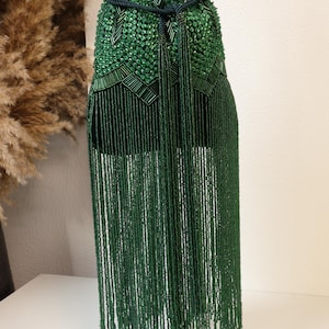 May include: A dark green bucket bag featuring an intricate exterior covered in faceted beads and long, dangling beaded fringe. The bag includes a dark green rope drawstring closure with two long tassels hanging from the center front.