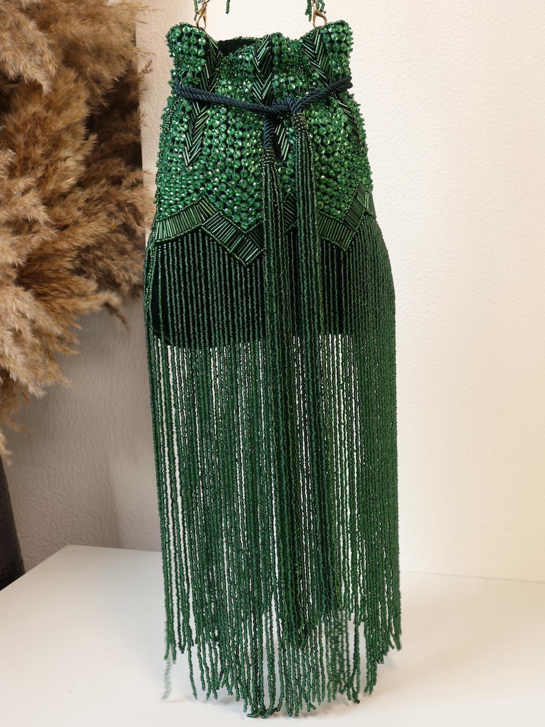 May include: A dark green bucket bag featuring an intricate exterior covered in faceted beads and long, dangling beaded fringe. The bag includes a dark green rope drawstring closure with two long tassels hanging from the center front.