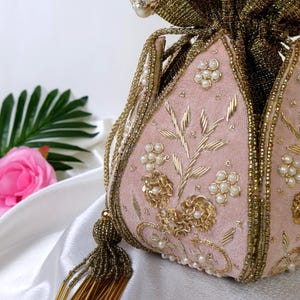 May include: A pink velvet drawstring bag with gold embroidery, pearl accents, and a beaded tassel. The bag features floral and leaf designs, with a gold fabric top and a pearl-beaded handle. A beautiful, decorative accessory.