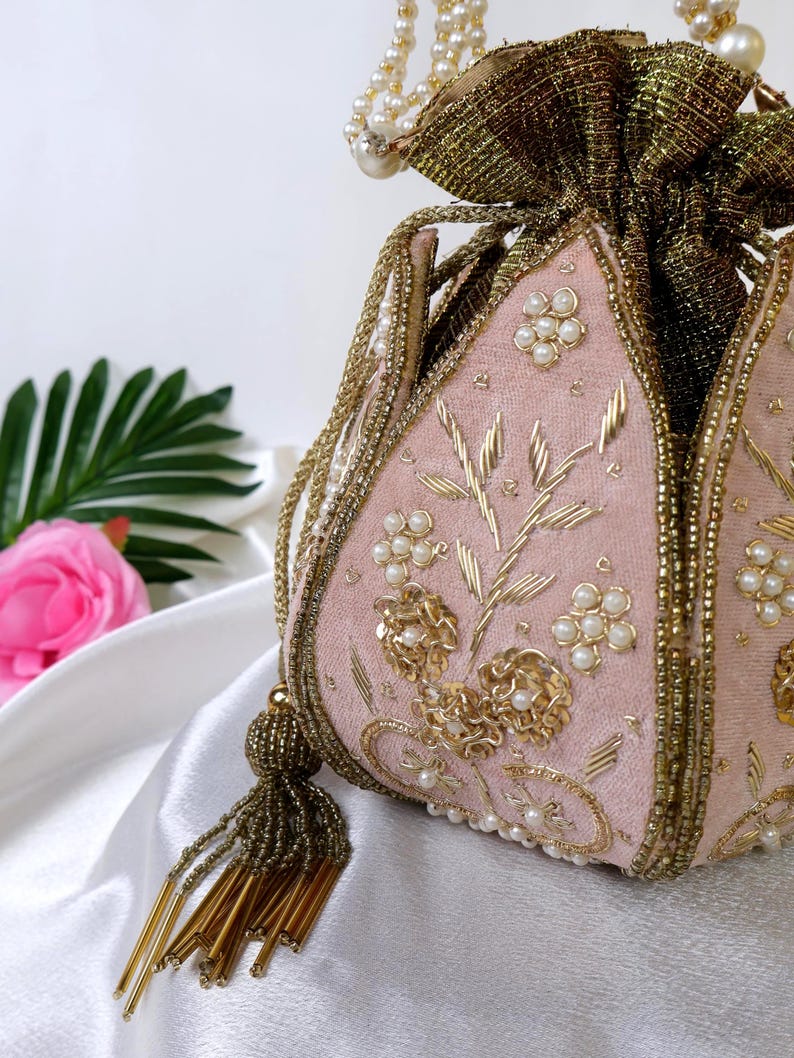 May include: A pink velvet drawstring bag with gold embroidery, pearl accents, and a beaded tassel. The bag features floral and leaf designs, with a gold fabric top and a pearl-beaded handle. A beautiful, decorative accessory.