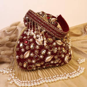 May include: A burgundy beaded clutch bag with a drawstring closure. The bag is adorned with pearls, gold beads, and decorative stones. The top of the bag has a burgundy velvet handle. Pearl and gold bead tassels hang from the bottom of the bag.