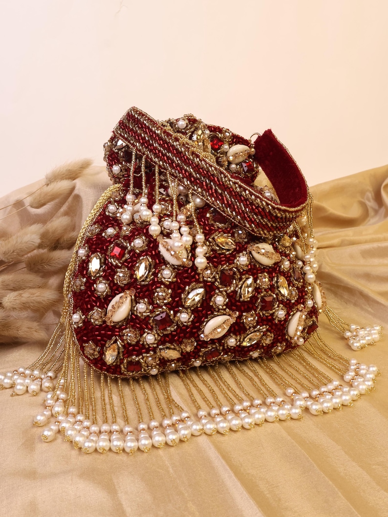 May include: A burgundy beaded clutch bag with a drawstring closure. The bag is adorned with pearls, gold beads, and decorative stones. The top of the bag has a burgundy velvet handle. Pearl and gold bead tassels hang from the bottom of the bag.