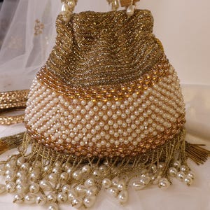May include: A gold beaded handbag with a pearl-like beaded bottom and fringe. The bag has a drawstring closure and pearl-like beaded handles. The bag is decorated with gold beads and a pearl-like beaded design.
