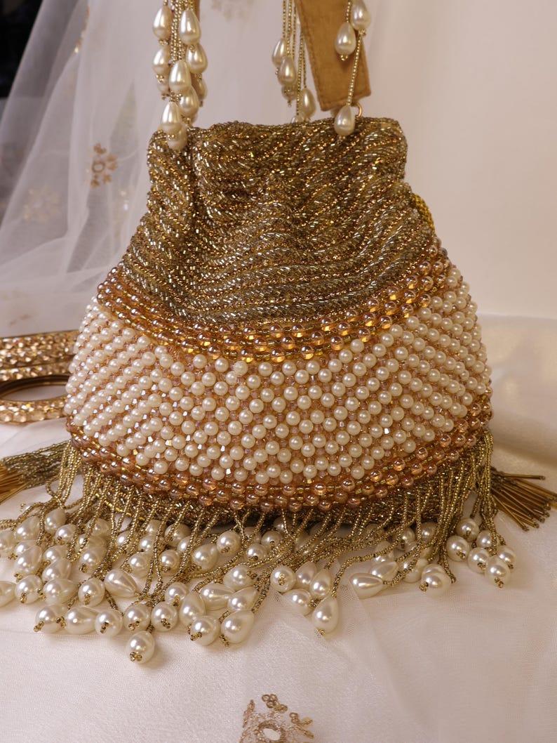 May include: A gold beaded handbag with a pearl-like beaded bottom and fringe. The bag has a drawstring closure and pearl-like beaded handles. The bag is decorated with gold beads and a pearl-like beaded design.