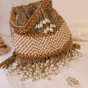 May include: A beaded pouch bag with a drawstring closure. The bag features a combination of gold and cream-colored beads, with pearl-like teardrop accents and gold fringe. The bag is likely used for carrying small items.