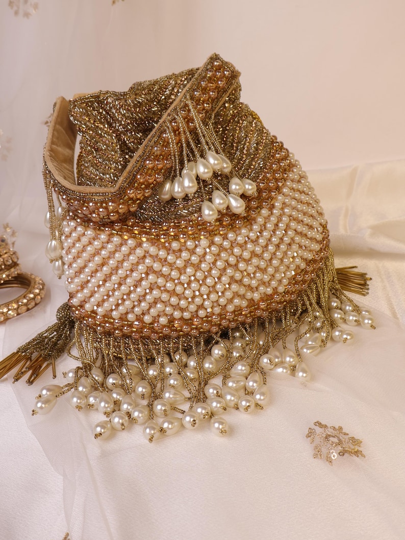 May include: A beaded pouch bag with a drawstring closure. The bag features a combination of gold and cream-colored beads, with pearl-like teardrop accents and gold fringe. The bag is likely used for carrying small items.