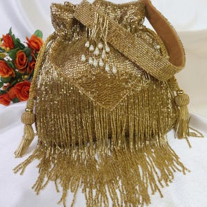 May include: A gold beaded drawstring bag with a matching shoulder strap. The bag features a square pattern and long fringe. The strap is lined with a brown material and adorned with white pearl-like beads. Tassels hang from the drawstring and the sides.