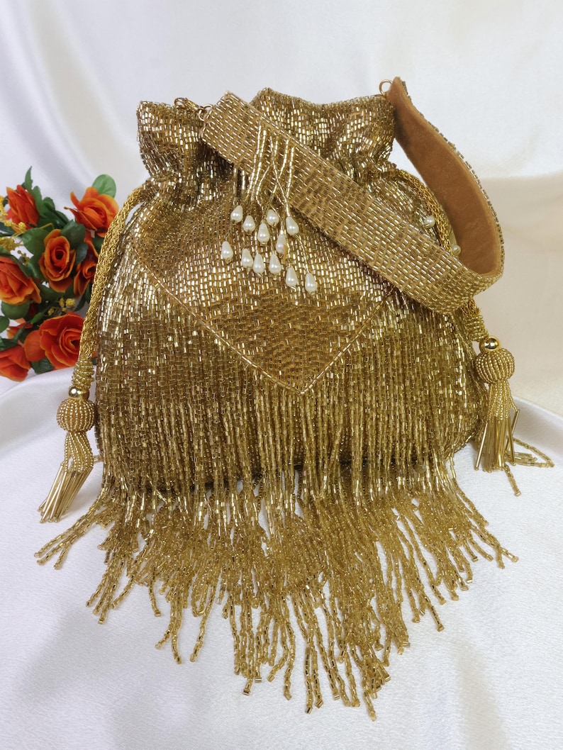 May include: A gold beaded drawstring bag with a matching shoulder strap. The bag features a square pattern and long fringe. The strap is lined with a brown material and adorned with white pearl-like beads. Tassels hang from the drawstring and the sides.