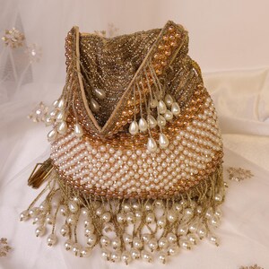 May include: A beaded pouch in gold and cream tones. The bag is covered in small, gold beads at the top, transitioning to cream-colored beads at the bottom. Cream-colored teardrop-shaped beads hang from the bottom edge. The bag has a drawstring closure.