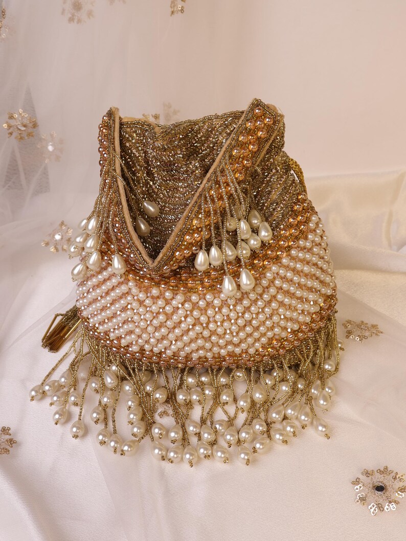 May include: A beaded pouch in gold and cream tones. The bag is covered in small, gold beads at the top, transitioning to cream-colored beads at the bottom. Cream-colored teardrop-shaped beads hang from the bottom edge. The bag has a drawstring closure.