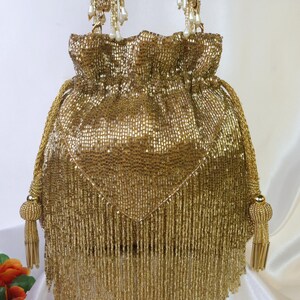 May include: A gold beaded drawstring bag with a fringe bottom and beaded tassels. The bag has a woven texture and a drawstring closure. The handles are made of gold rope and adorned with pearl-like beads.