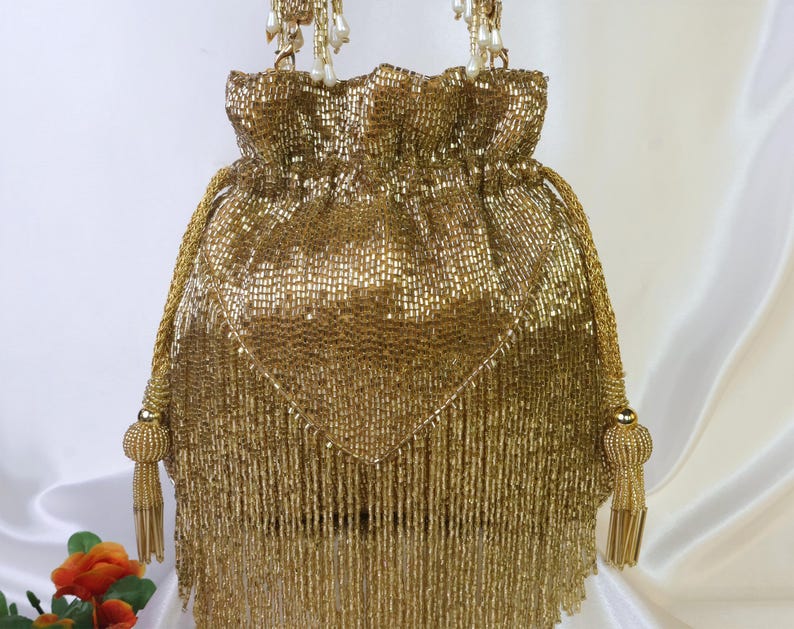 May include: A gold beaded drawstring bag with a fringe bottom and beaded tassels. The bag has a woven texture and a drawstring closure. The handles are made of gold rope and adorned with pearl-like beads.
