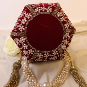 May include: A red velvet clutch purse with gold and pearl embellishments. The purse has a hexagonal shape and a tassel closure.