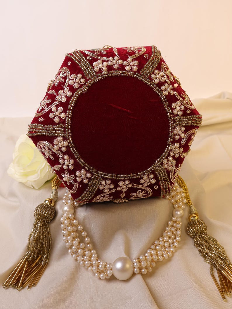 May include: A red velvet clutch purse with gold and pearl embellishments. The purse has a hexagonal shape and a tassel closure.