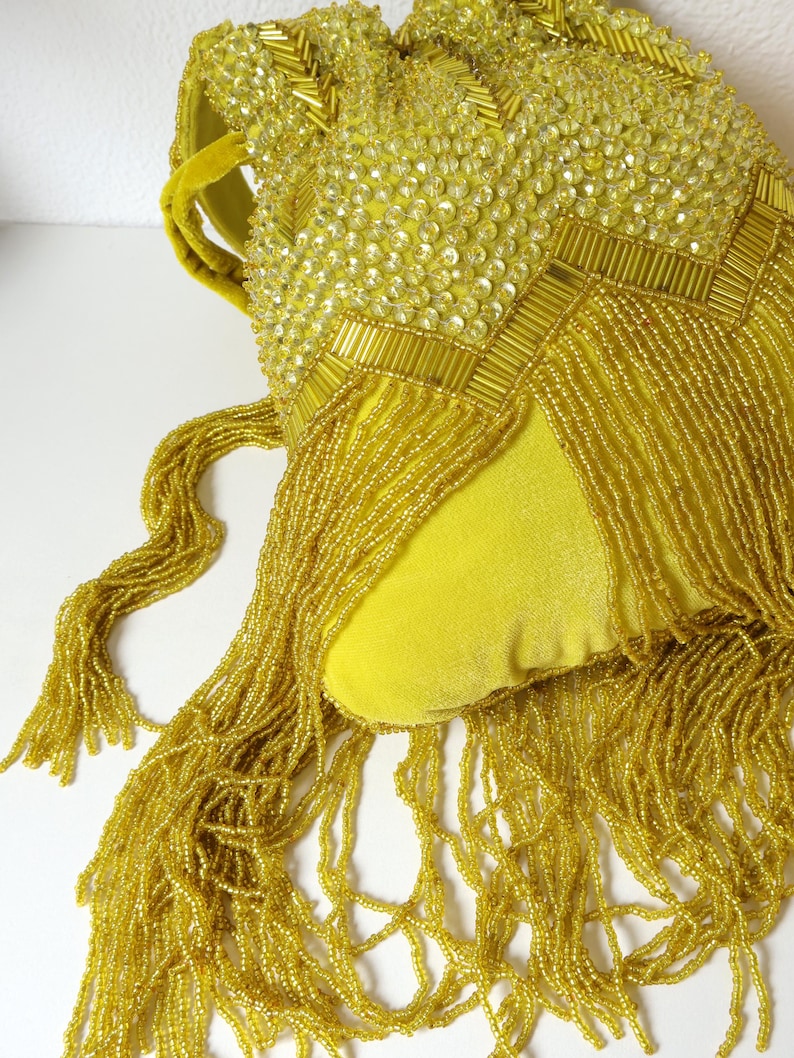 May include: A bright yellow velvet handbag featuring intricate beadwork and sequins in a chevron pattern. The bag is adorned with long, cascading yellow beaded fringe that hangs from the bottom, and it includes a matching yellow velvet handle.