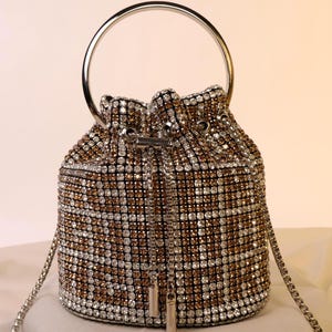Può includere: Una piccola e glamour borsa a secchiello ricoperta di strass scintillanti in un motivo a scacchi argento e oro. È dotata di un manico in metallo argentato e di una delicata tracolla a catena. La borsa ha una chiusura con coulisse e una cerniera argentata.