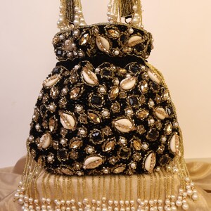 May include: A black, beaded handbag with gold and pearl accents. The bag features a velvet texture, adorned with gold-toned embellishments, white shells, and small pearls. Gold beaded fringe and handles add to the bag's ornate design. The bag is a stylish accessory.