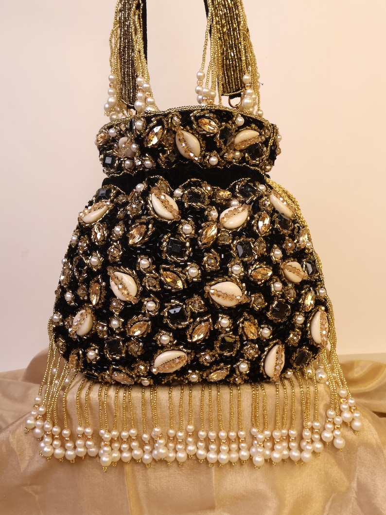 May include: A black, beaded handbag with gold and pearl accents. The bag features a velvet texture, adorned with gold-toned embellishments, white shells, and small pearls. Gold beaded fringe and handles add to the bag's ornate design. The bag is a stylish accessory.