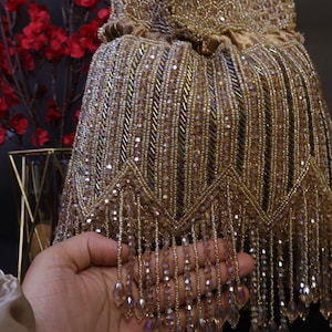 May include: A gold beaded handbag with a drawstring closure. The bag is decorated with rows of small beads and dangling fringe. The bag is held in front of a dark background with red flowers.