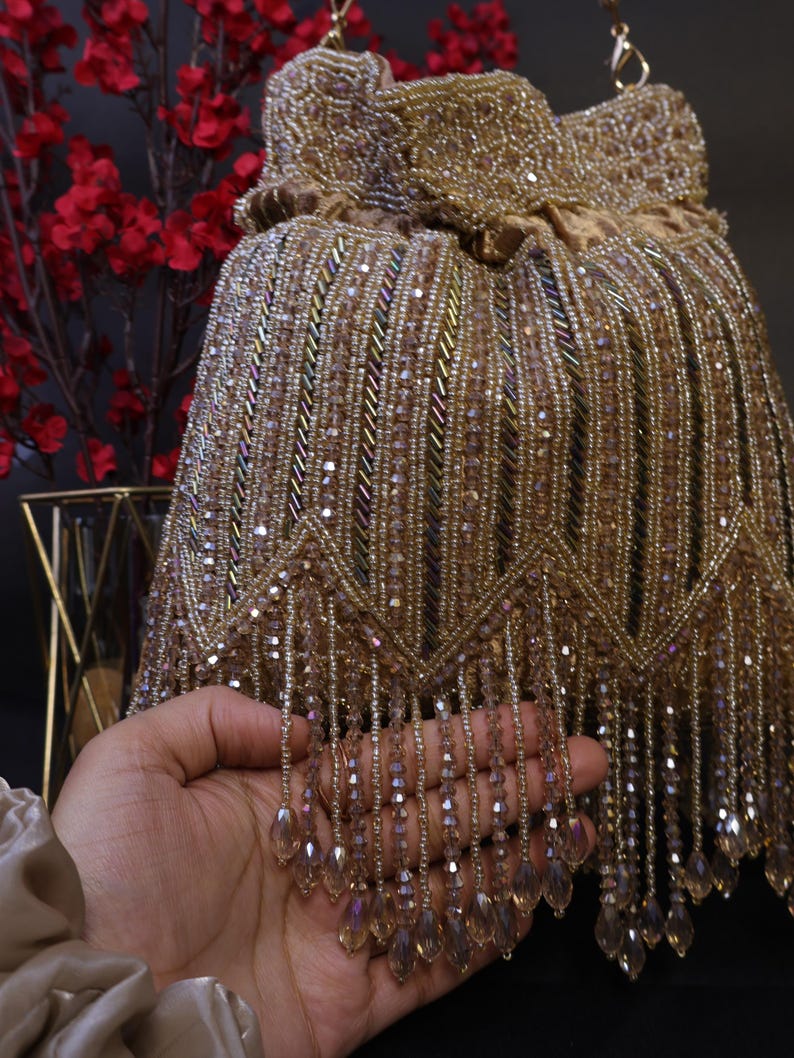 May include: A gold beaded handbag with a drawstring closure. The bag is decorated with rows of small beads and dangling fringe. The bag is held in front of a dark background with red flowers.