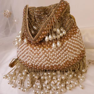 May include: A gold beaded drawstring bag with white pearl accents. The bag features a gold beaded top and a body covered in white pearls. Long pearl and gold bead fringe hangs from the bottom. The bag is a formal accessory.