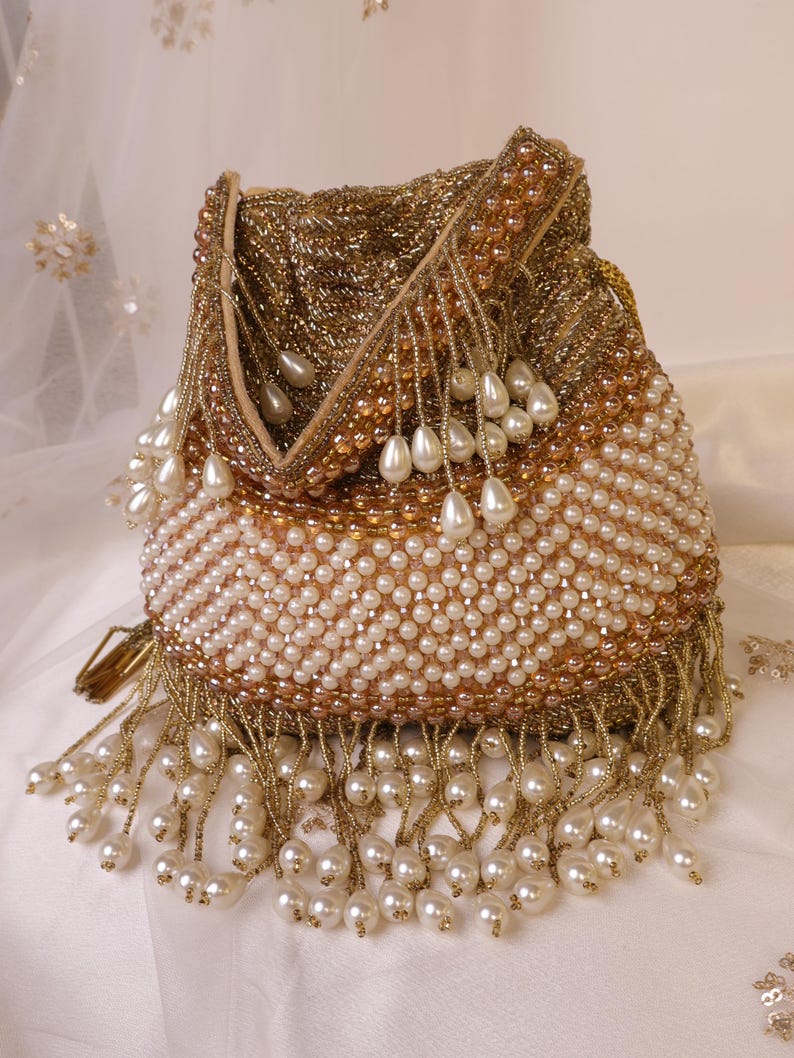May include: A gold beaded drawstring bag with white pearl accents. The bag features a gold beaded top and a body covered in white pearls. Long pearl and gold bead fringe hangs from the bottom. The bag is a formal accessory.