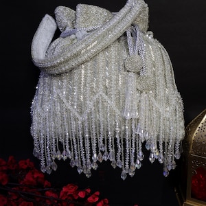 May include: A white beaded bag with a drawstring closure and a long, beaded handle. The bag is heavily embellished with silver beads and dangling crystal accents, creating a glamorous, elegant look. The bag is set against a dark background.