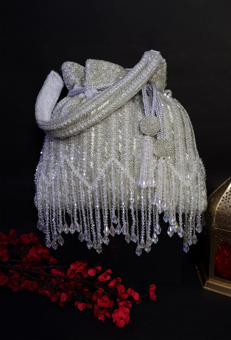 May include: A white beaded bag with a drawstring closure and a long, beaded handle. The bag is heavily embellished with silver beads and dangling crystal accents, creating a glamorous, elegant look. The bag is set against a dark background.