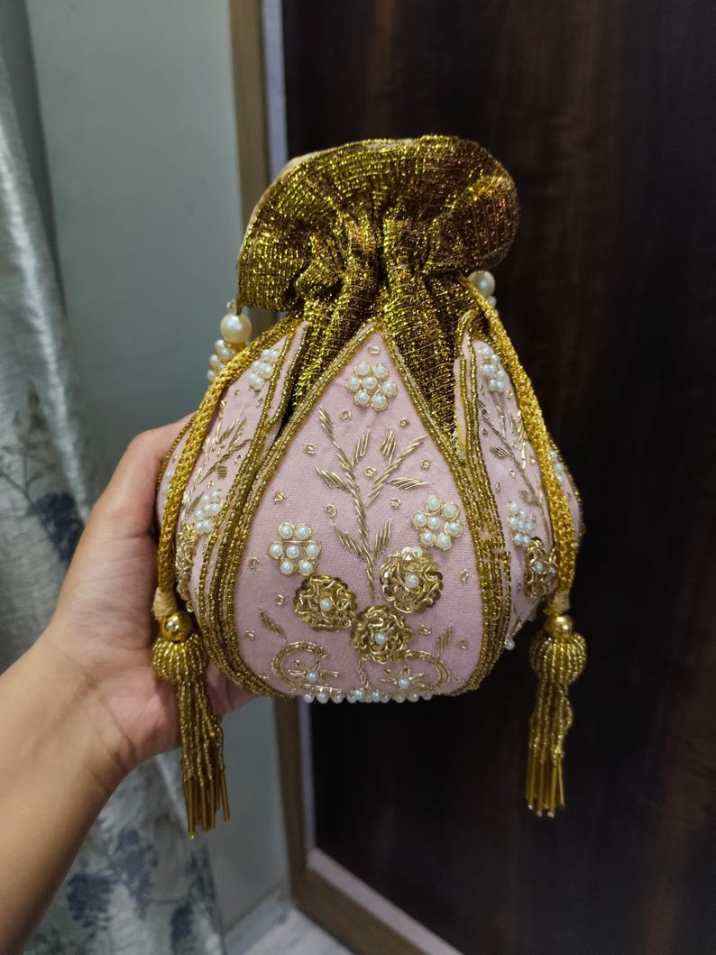 May include: A pink drawstring pouch with gold embroidery and pearl accents. The pouch features floral and leaf designs, gold tassels, and a gold mesh top. The bag is likely used for carrying small items.