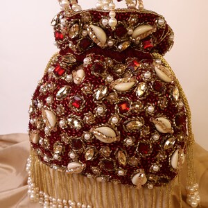 May include: A burgundy beaded handbag with a drawstring closure, adorned with pearls, gold beads, and gemstones. The bag features a fringe of gold beads and a handle embellished with pearls. The bag is approximately 8 inches tall.