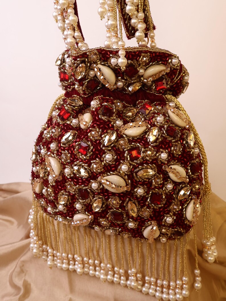 May include: A burgundy beaded handbag with a drawstring closure, adorned with pearls, gold beads, and gemstones. The bag features a fringe of gold beads and a handle embellished with pearls. The bag is approximately 8 inches tall.
