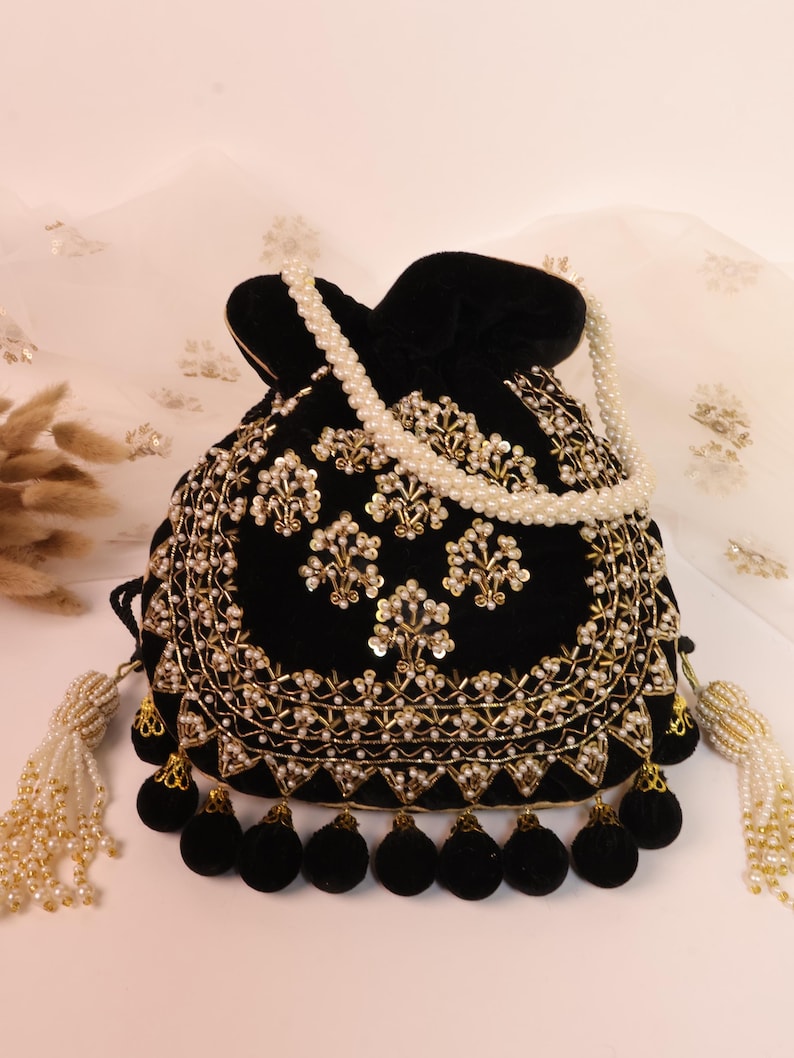 May include: A black velvet drawstring bag with gold embroidery and pearl detailing. The bag has a beaded pearl strap and tassels with black pom-poms. The design is ornate and elegant, suitable for special occasions.