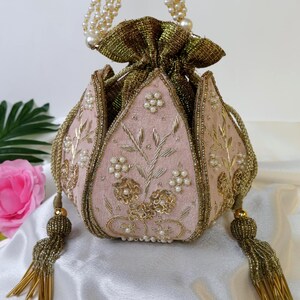 May include: A pink and gold beaded clutch bag with a floral design and pearl accents. The bag has a drawstring closure and a beaded chain strap.