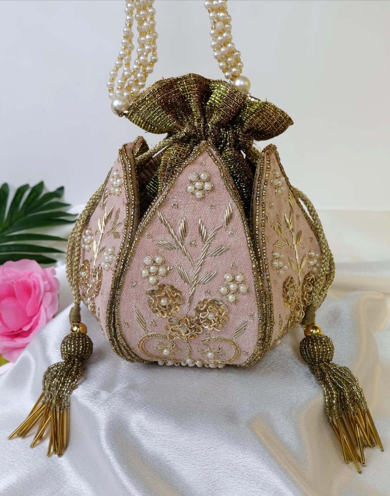 May include: A pink and gold beaded clutch bag with a floral design and pearl accents. The bag has a drawstring closure and a beaded chain strap.