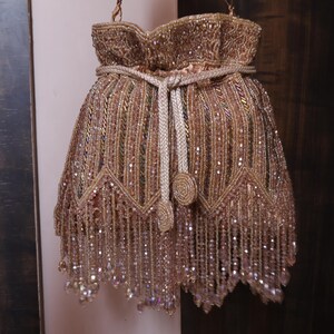 May include: A gold and brown beaded drawstring bag. The bag has a fringed bottom and a braided drawstring closure. The design features intricate beadwork and a brown strap. The bag is decorated with a detailed pattern.