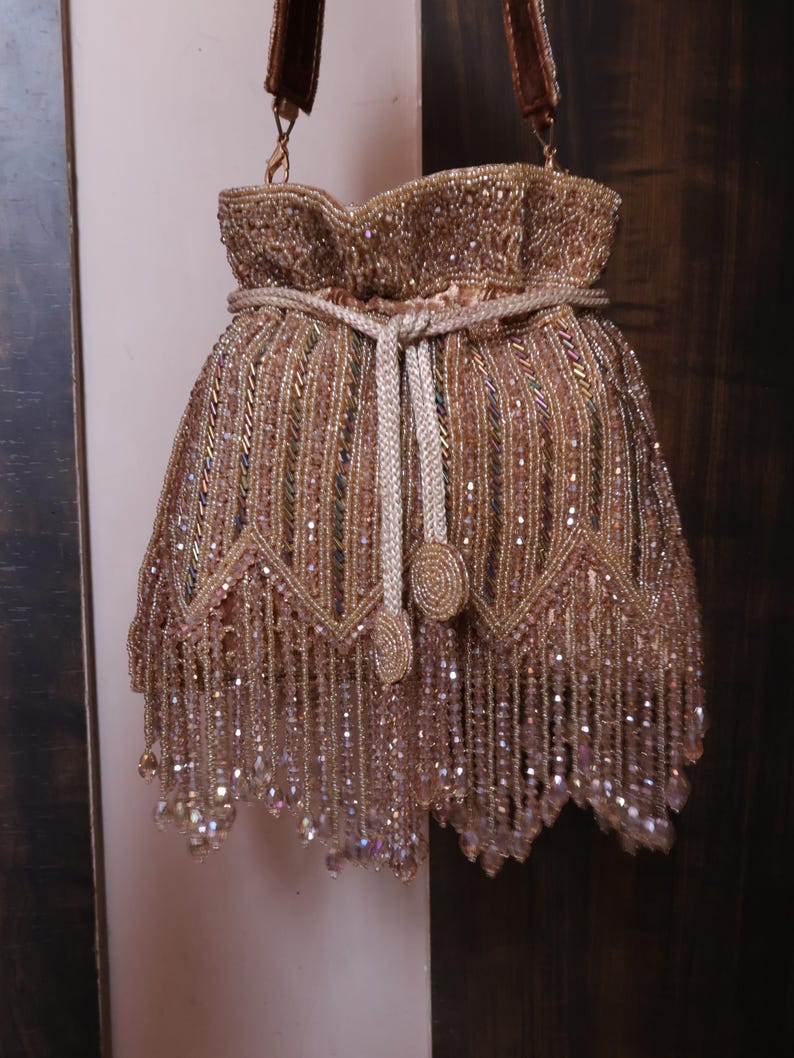May include: A gold and brown beaded drawstring bag. The bag has a fringed bottom and a braided drawstring closure. The design features intricate beadwork and a brown strap. The bag is decorated with a detailed pattern.