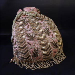 Puede incluir: Una bolsa de cordón rosa con adornos dorados y blancos. La bolsa tiene una franja de cuentas y un cierre de cordón.