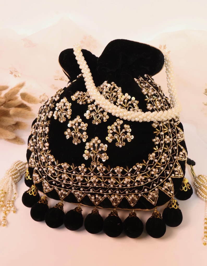 May include: Black velvet pouch bag with gold and pearl detailing. The bag has a drawstring closure, black pom-pom fringe, and a pearl beaded handle. A decorative accessory for special events.