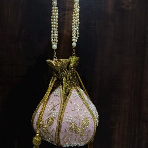 May include: A pink embroidered pouch bag with gold accents and a pearl beaded handle. The bag features floral embroidery and a gold tassel. The bag is held up by a pearl beaded handle.