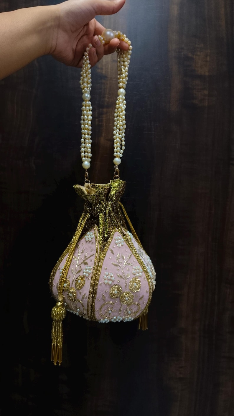 May include: A pink embroidered pouch bag with gold accents and a pearl beaded handle. The bag features floral embroidery and a gold tassel. The bag is held up by a pearl beaded handle.
