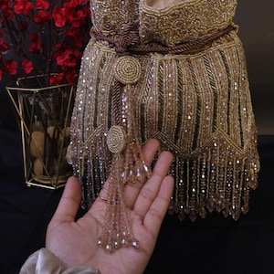 May include: A gold beaded handbag with a drawstring closure and a long shoulder strap. The bag features intricate beadwork and a tassel detail. The bag is approximately 20 cm tall and 18 cm wide.