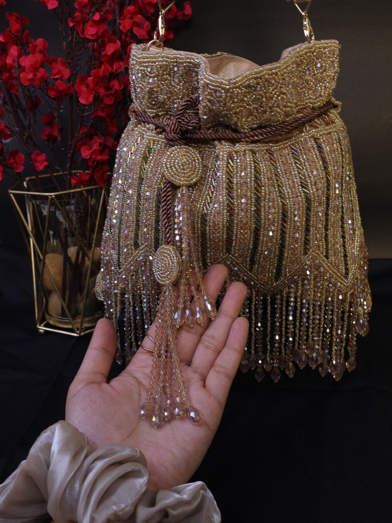 May include: A gold beaded handbag with a drawstring closure and a long shoulder strap. The bag features intricate beadwork and a tassel detail. The bag is approximately 20 cm tall and 18 cm wide.