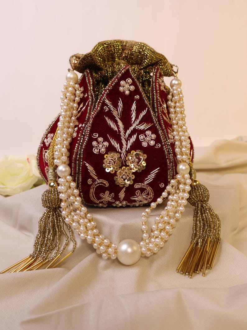 May include: A red velvet drawstring bag with gold and silver embroidery and embellishments. The bag has a long pearl and gold beaded strap with a large pearl accent.