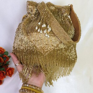 May include: A gold beaded drawstring bag with a matching strap and beaded fringe. The bag is embellished with small white pearl-like beads. A hand wearing gold bangles and a ring holds the bag.