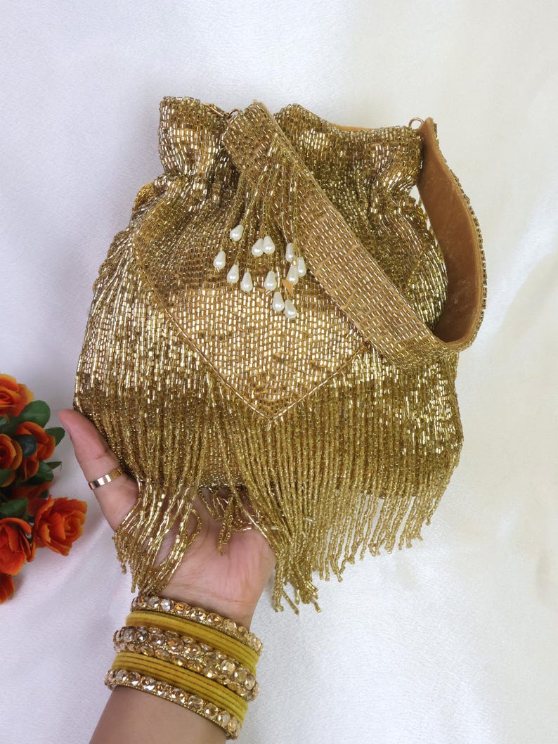 May include: A gold beaded drawstring bag with a matching strap and beaded fringe. The bag is embellished with small white pearl-like beads. A hand wearing gold bangles and a ring holds the bag.